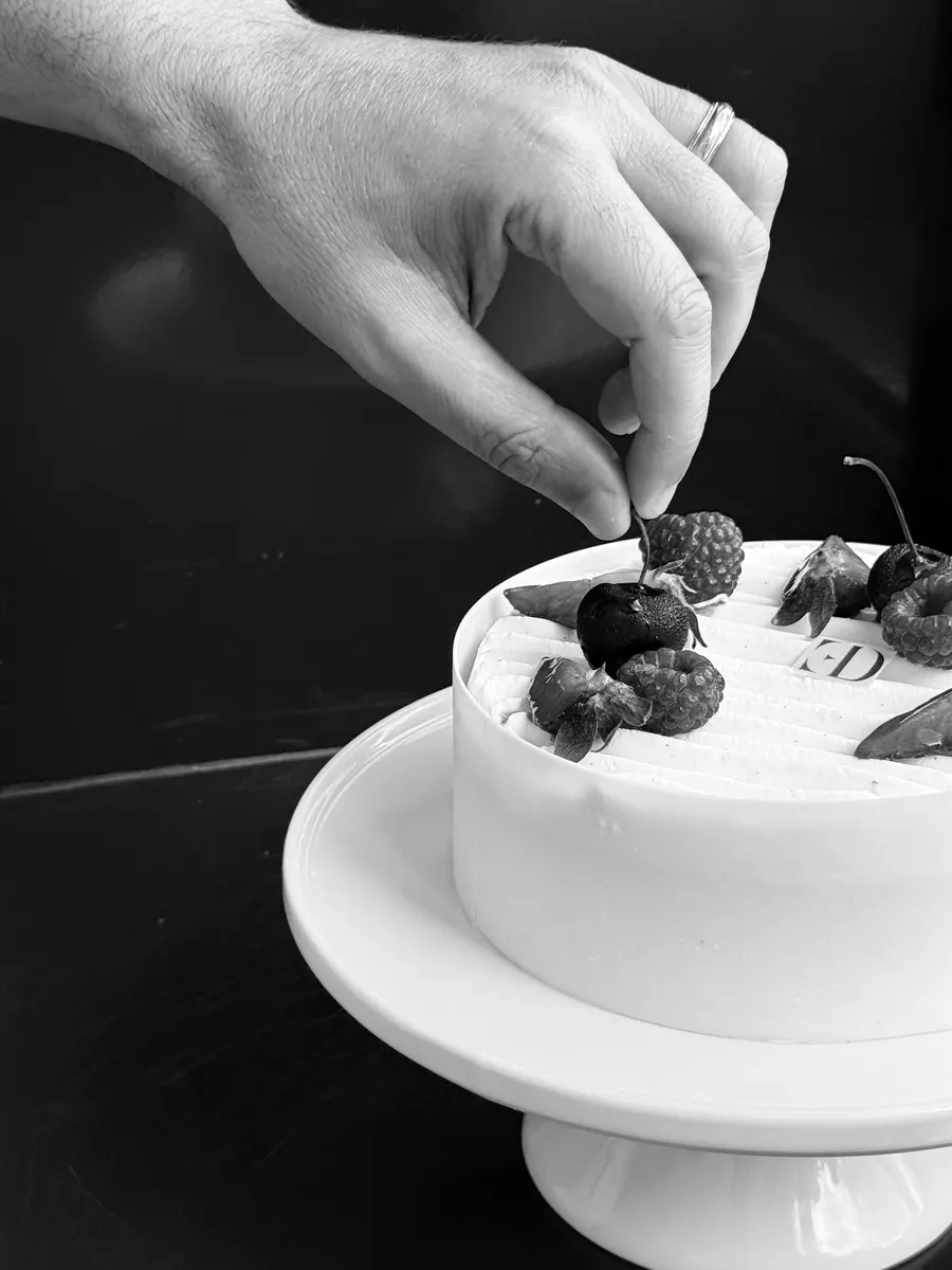 Ali's hands placing fresh berries on a white-frosted entremet, photographed black and white.