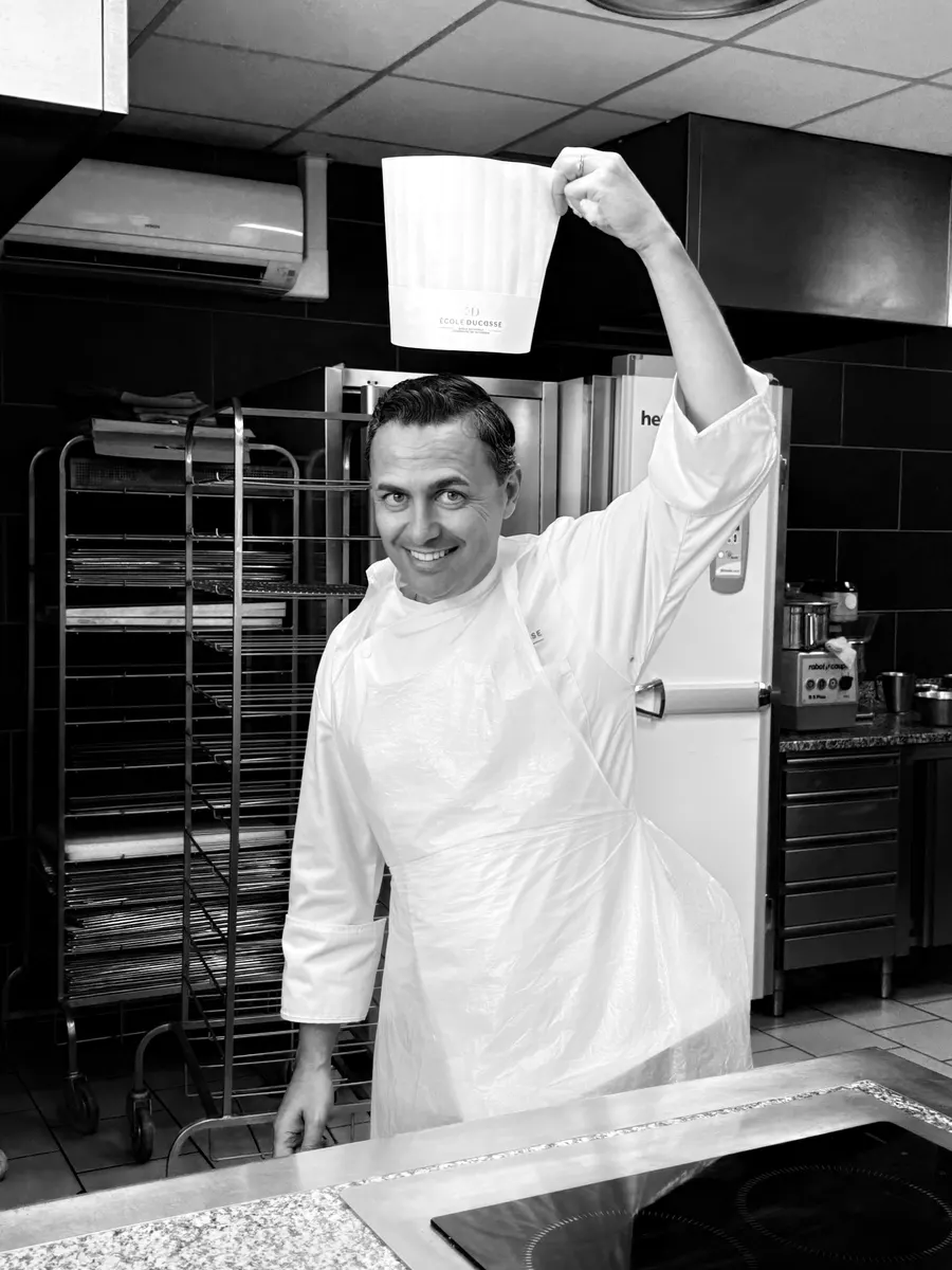 Ali Al Ghoul in chef whites holding up his École Ducasse toque in the kitchen — black and white.