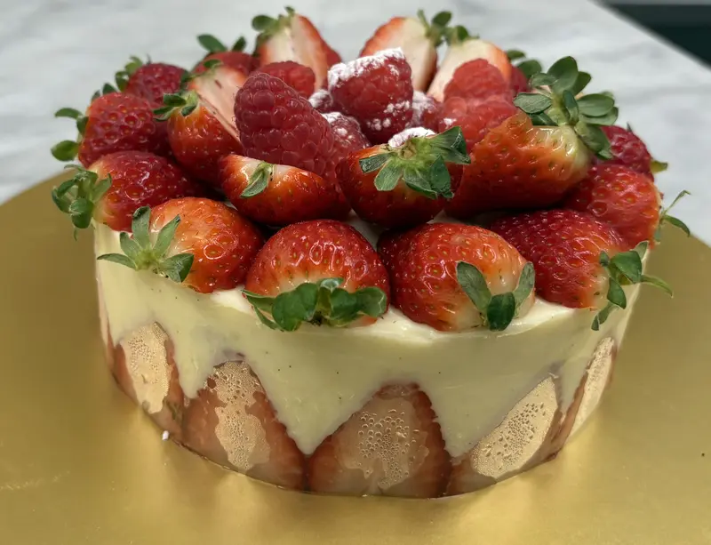 Ali's traditional Fraisier — vanilla génoise layered with crème mousseline, walls lined with halved fresh strawberries showing through the sides, crowned with whole strawberries and raspberries dusted with sugar.