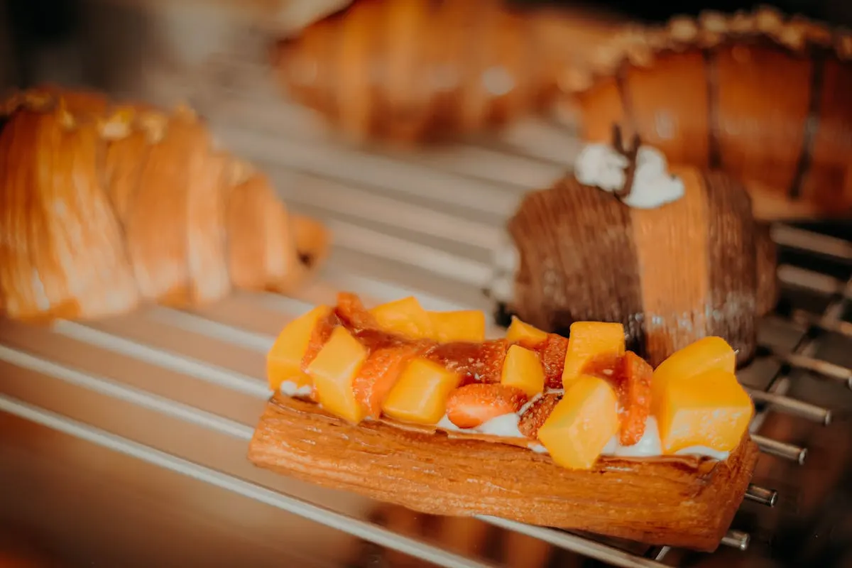 A puff-pastry mille-feuille topped with cubed mango and strawberry on a cooling rack beside croissants.