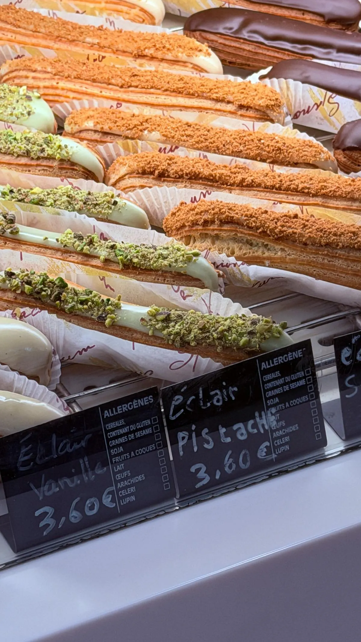A row of vanilla and pistachio éclairs in a Paris bakery display case with handwritten price cards.