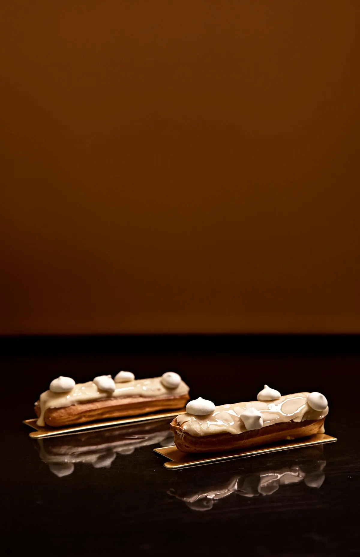 Two cream-glazed éclairs topped with meringue dots on a dark reflective surface.