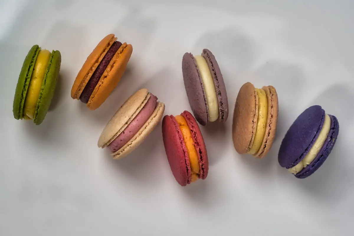 A row of eight French macarons in green, orange, lavender, beige, red, light brown, and purple.