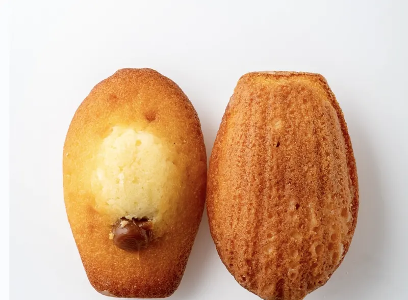 A scallop-shape madeleine with a salted-caramel dot on top beside a cut half showing the golden crumb.