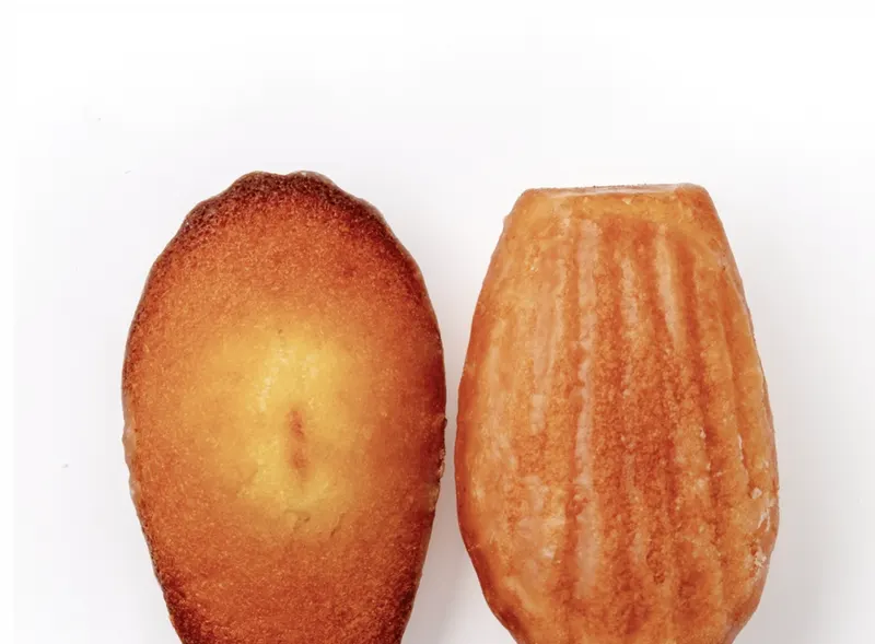 A scallop-shape lemon-glazed madeleine and a cut half on white, showing a soft buttery crumb.