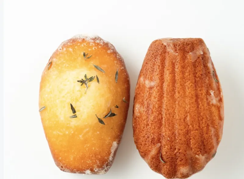 A scallop-shape madeleine glazed with lime and dotted with fresh thyme leaves, beside a cut half showing a tender pale-green-flecked crumb.