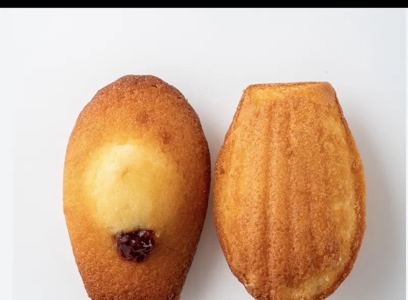 A scallop-shape madeleine with a pink raspberry-jam dot on top beside a cut half showing the buttery crumb.