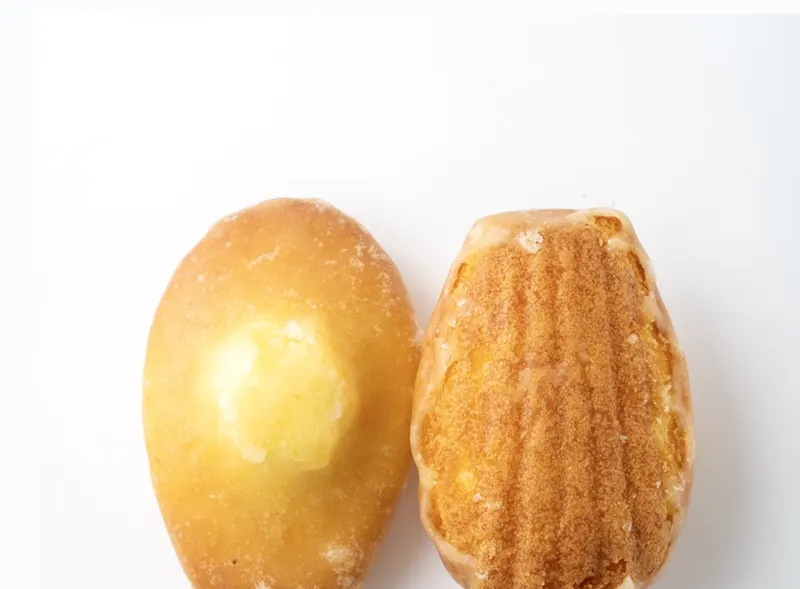 A scallop-shape madeleine with a glossy orange glaze beside a cut half showing the warm amber crumb.