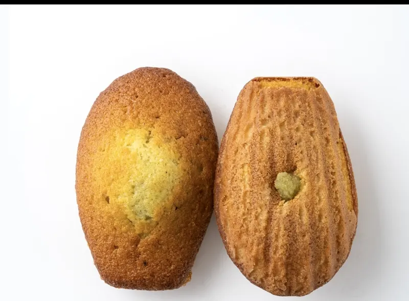 A scallop-shape pistachio madeleine and a cut half showing the green pistachio-flecked crumb.