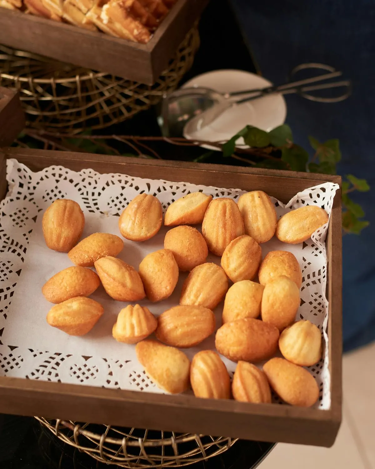 A wooden serving tray full of golden madeleines on a lace doily, photographed from above against deep navy linen.