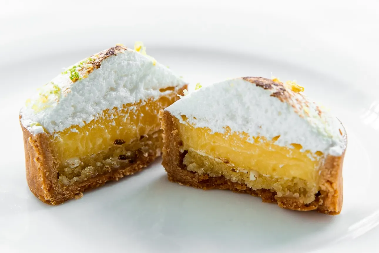 The same lemon tart cut in half, showing the pale almond layer at the base, a thick band of lemon curd above it, and the meringue on top.