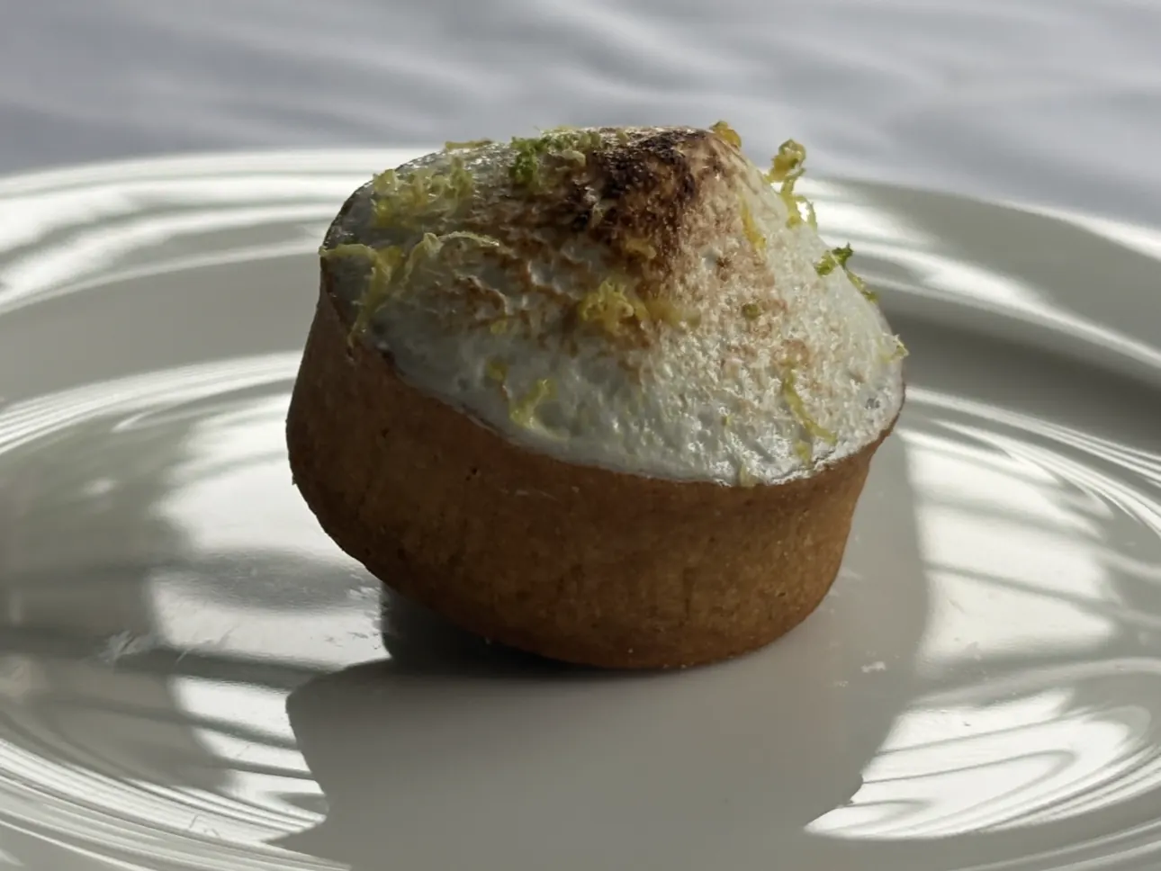 The lemon tart photographed in daylight on a white plate, the torched meringue and zest lit from the side.