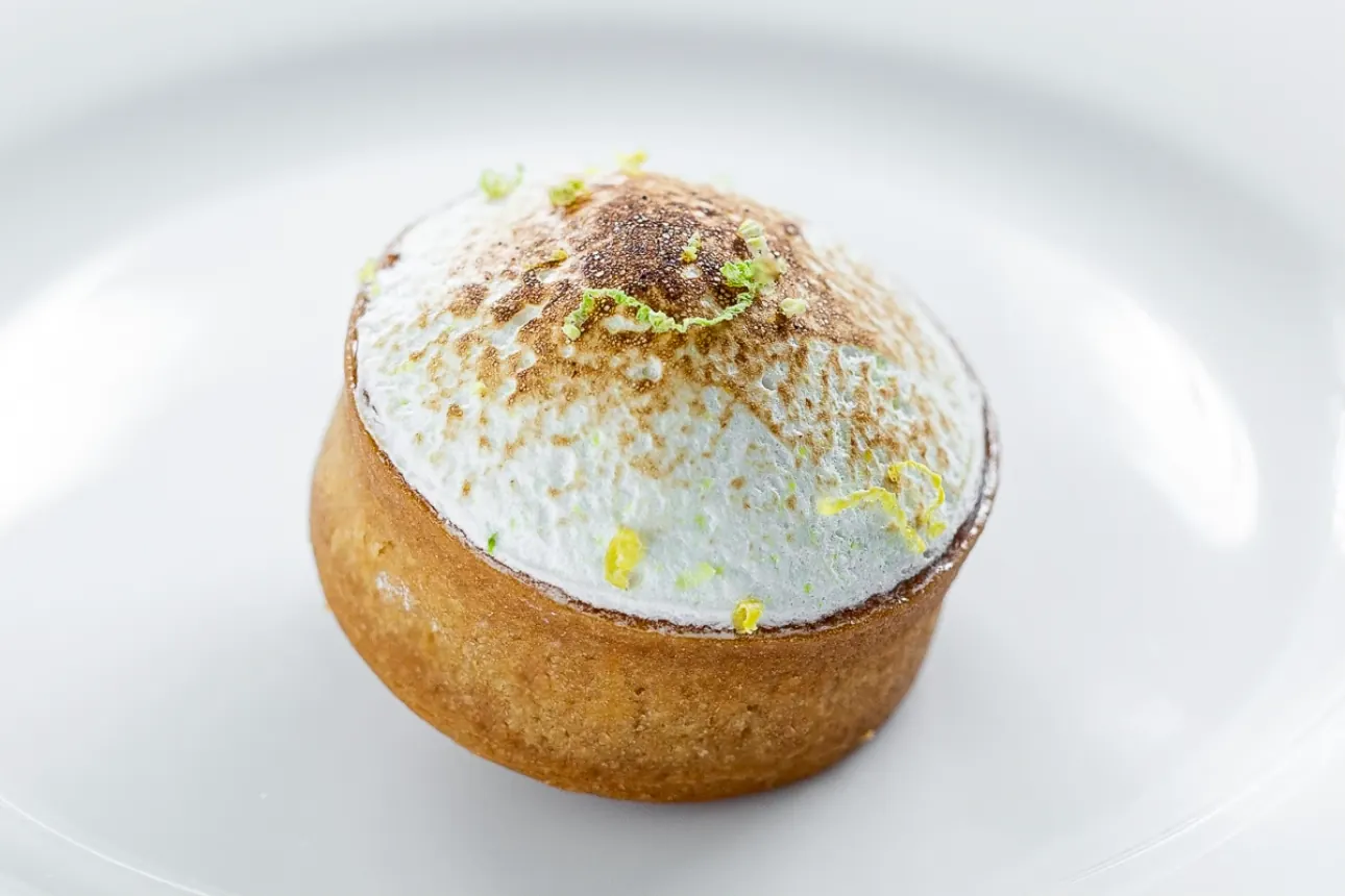 A lemon tart with a domed cap of Italian meringue torched to a soft gold, scattered with lemon and lime zest, on a white plate.