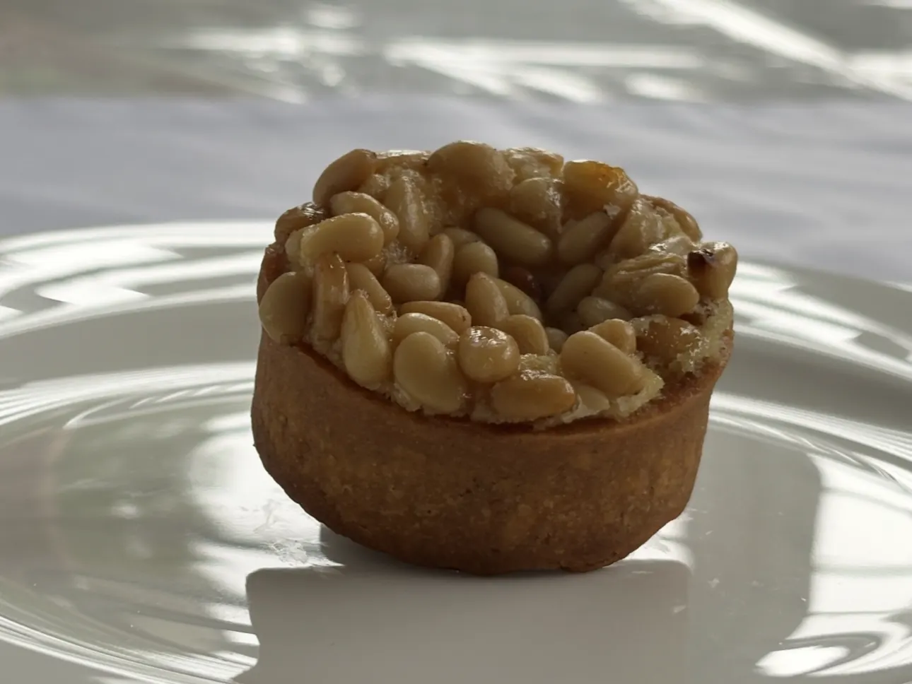 The pine-nut tart photographed in daylight on a white plate, the glazed nuts catching the sun.