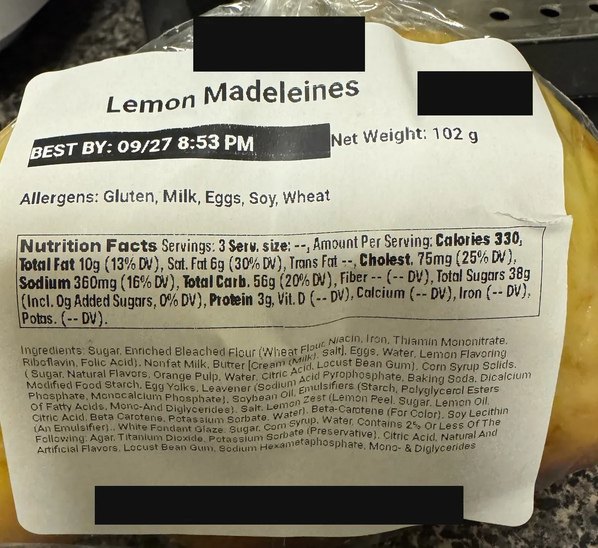 The printed label on a bag of lemon madeleines from a national café chain, with the brand name, price and maker's address blacked out. The ingredient list runs eight dense lines and includes soybean oil, titanium dioxide and sodium hexametaphosphate.
